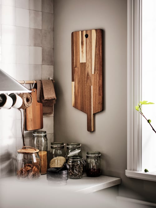 A brown chopping board hangs from a grey wall. Under the board are multiple jars with lid, all on a countertop.