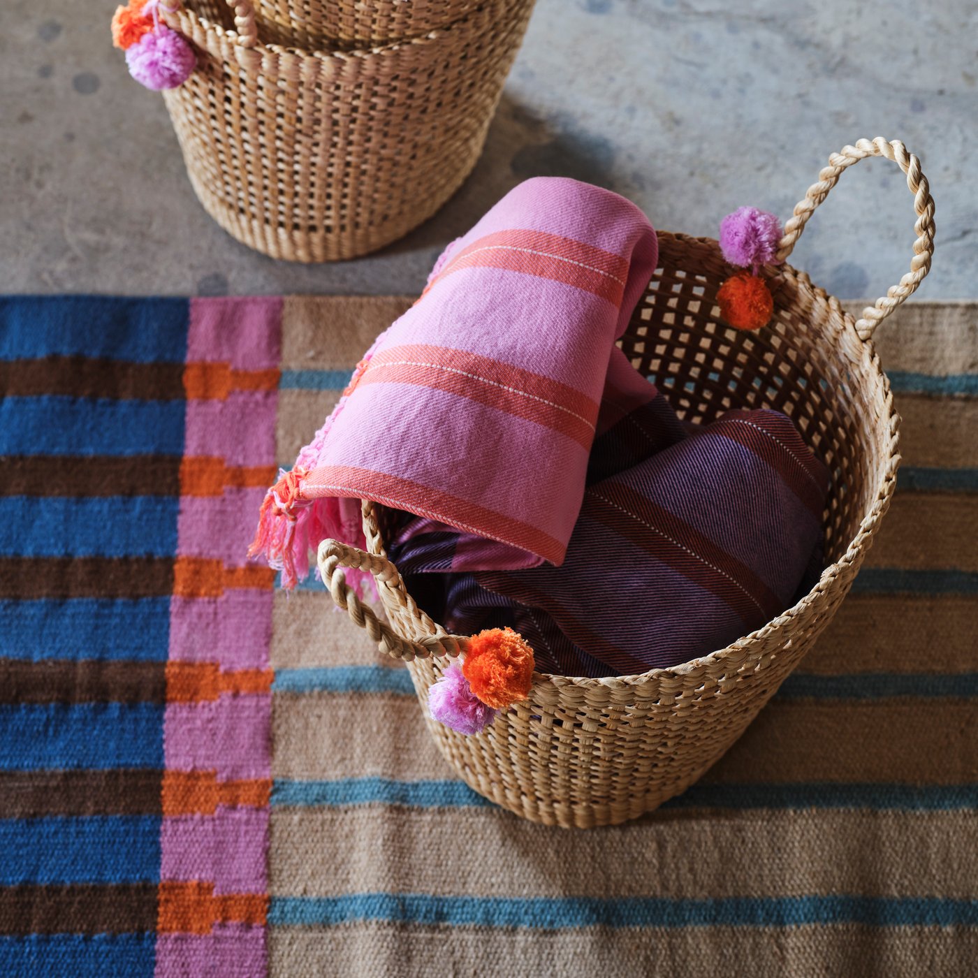 A couple of MÄVINN throws in pink and orange lie in a MÄVINN basket made of banana fibres with small pompoms on the handles.