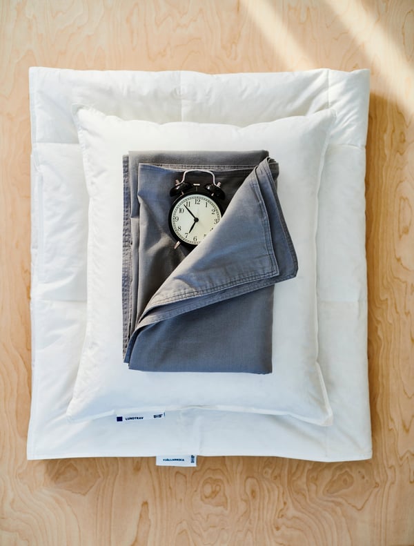 FJÄLLARNIKA light warm duvet folded neatly on the floor with a LUNDTRAV pillow and ÄNGSLILJA grey duvet cover placed on top with a DEKAD alarm clock.  