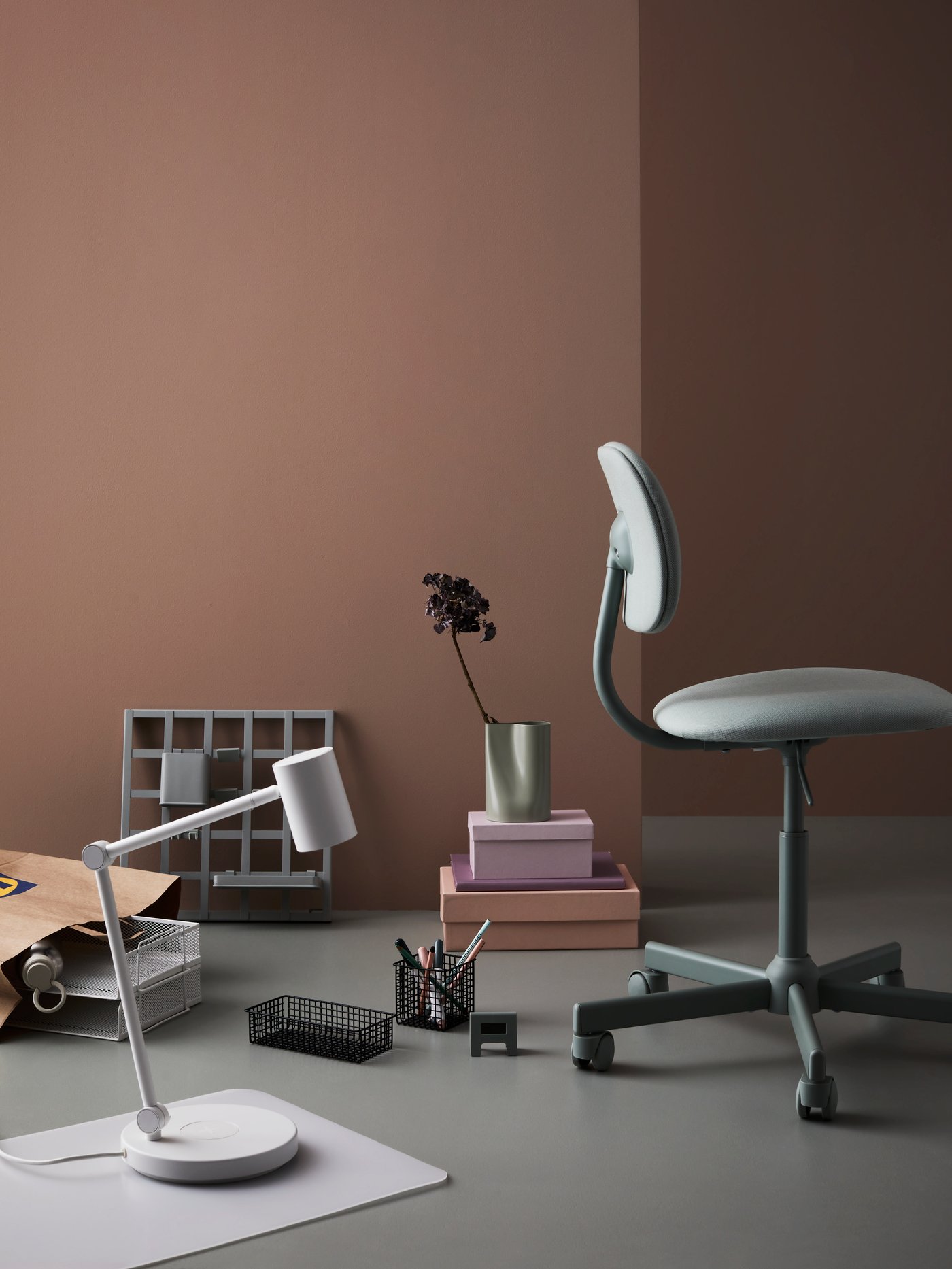 Schreibtischutensilien auf dem Fussboden, u. a. mit PLUGGLAND Drahtkörben in Nähe eines BLECKBERGET Drehstuhls. In der Nähe steht eine NYMÅNE Leuchte.