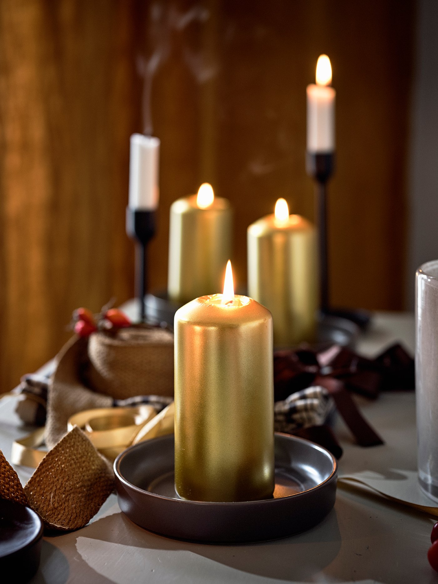 Gold FENOMEN candles sit on a table, in the back ground tall candles can be seen in black candle holders. 