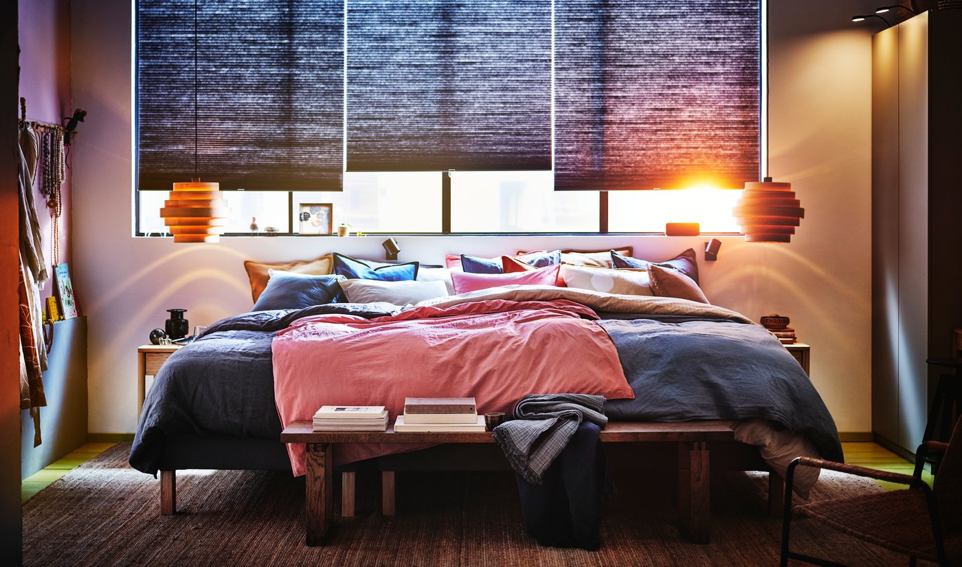 Soveværelse med bred seng foran et vindue. Næsten nedrullede gardiner, hvor den orange sol kigger ind.