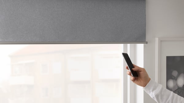 La imagen muestra una mano con un smartphone apuntando a una cortina roller eléctrica FYRTUR de color gris, a medio subir. 