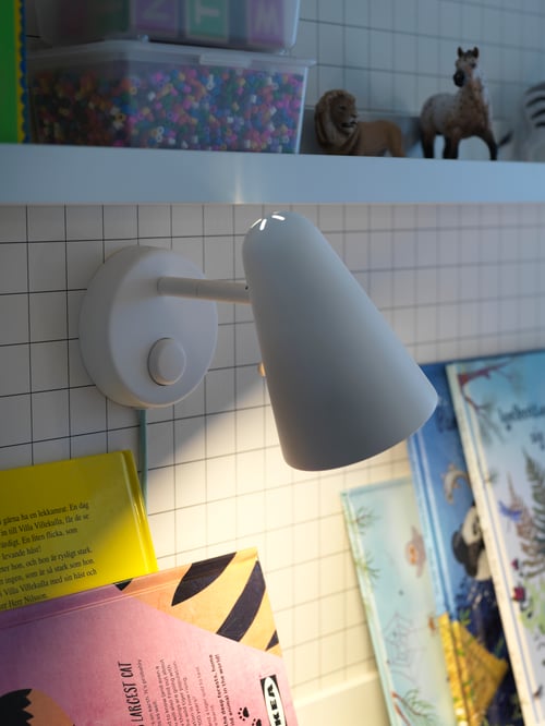 Une lampe pour enfants blanche sur un mur au-dessus de plusieurs livres et sous une étagère contenant une boîte de perles et des animaux-jouets.