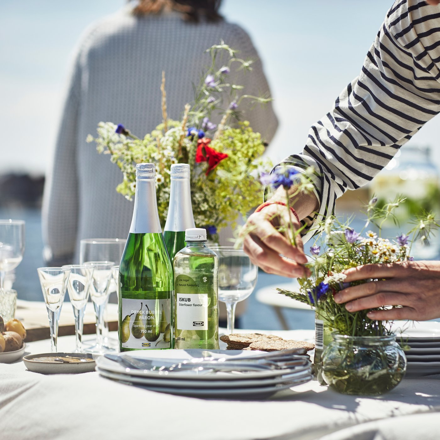 En person er ved at dække et bord til midsommer med en hvid dug, flasker og forskelligt service i midten af bordet står en buket med vilde blomster.