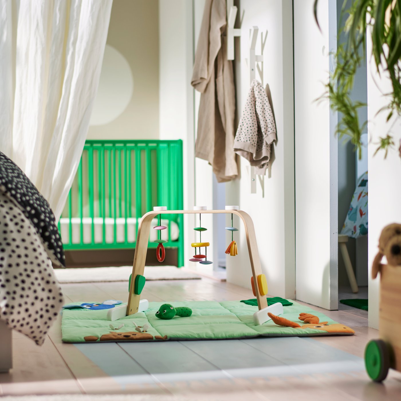 Ein buntes LEKA-Babygymnastikgerät mit hängenden Spielzeugen auf einer grünen Spielmatte auf dem Boden in einem Schlafzimmer mit einem grünen GULLIVER-Kinderbett.