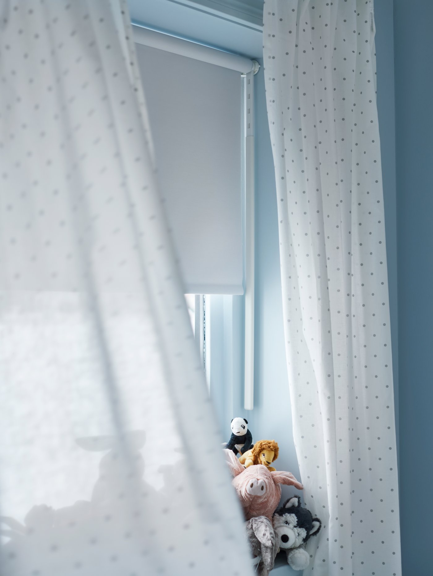 Una ventana con peluches en el alféizar, cortinas LEN blancas con puntos y un estor FRIDANS blanco.