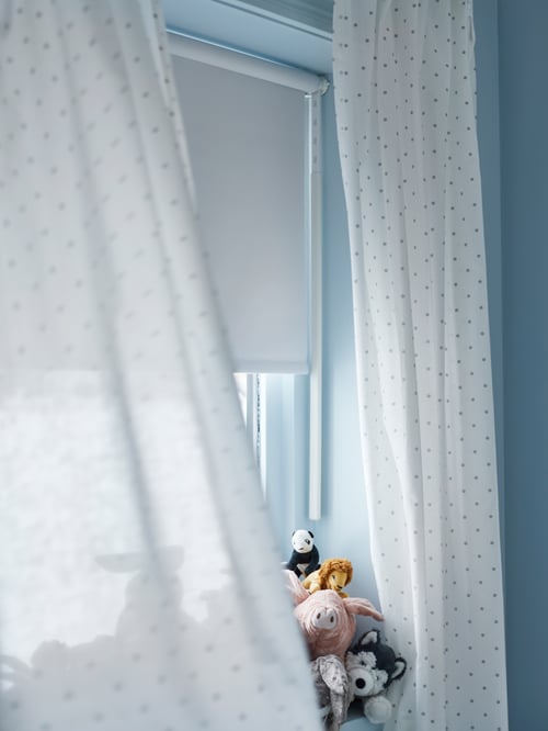 A window with soft toys on the window sill, dotted white LEN curtains and a white FRIDANS block-out roller blind.