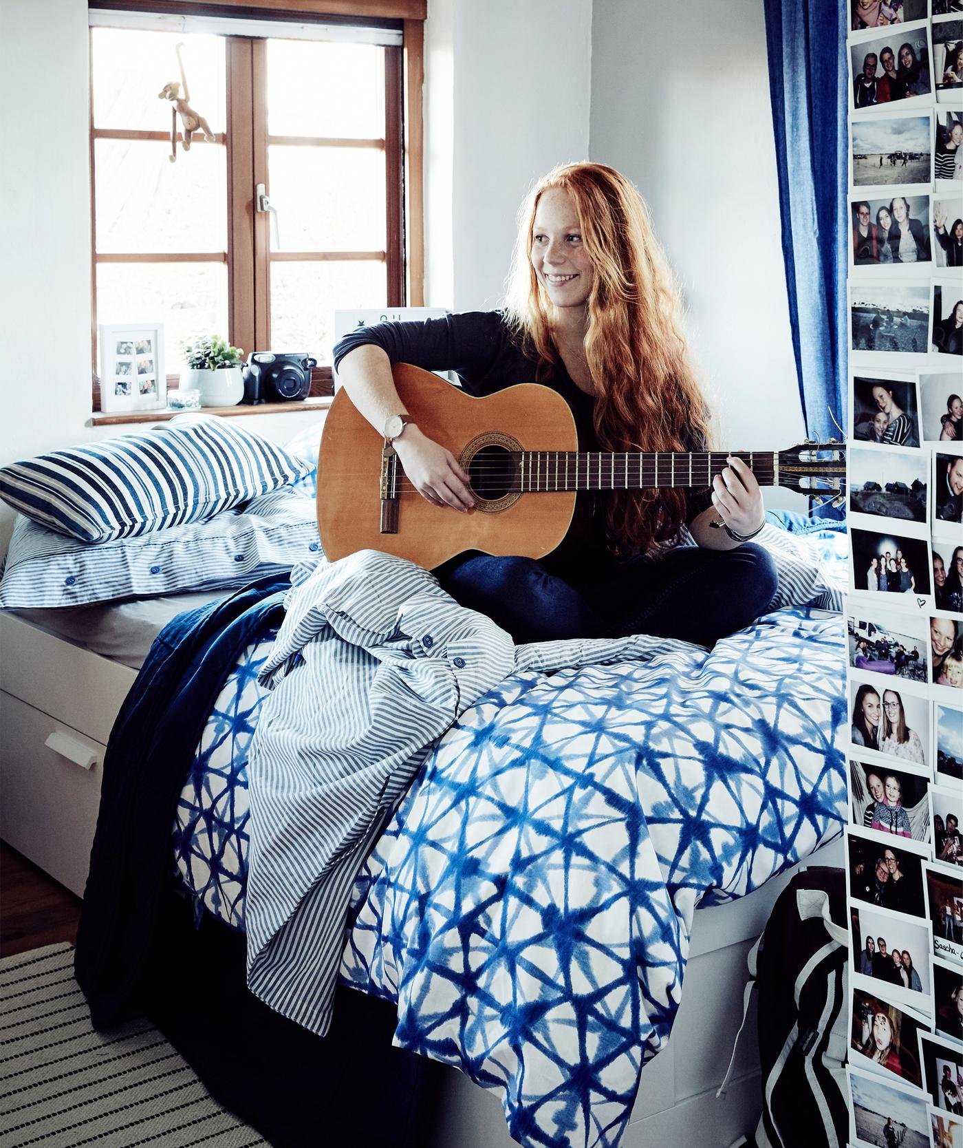 Ein Foto von Stine, die auf ihrem Bett Gitarre spielt, unter ihr STJÄRNFLOCKA Bettwäsche-Set in Weiss/Blau.