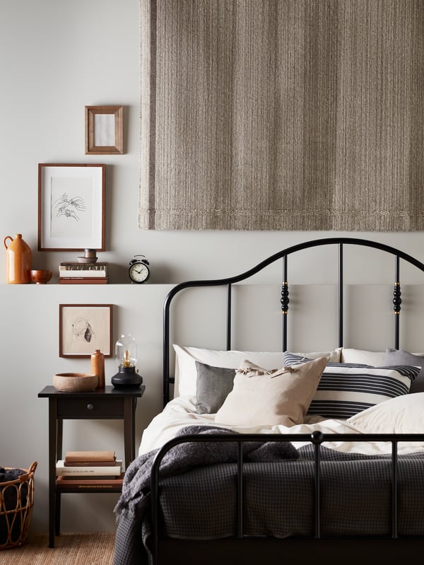 A bedroom with a black SAGSTUA bed made with grey-beige ÄNGSLILJA bed linen, a rug on the wall and a HEMNES bedside table.