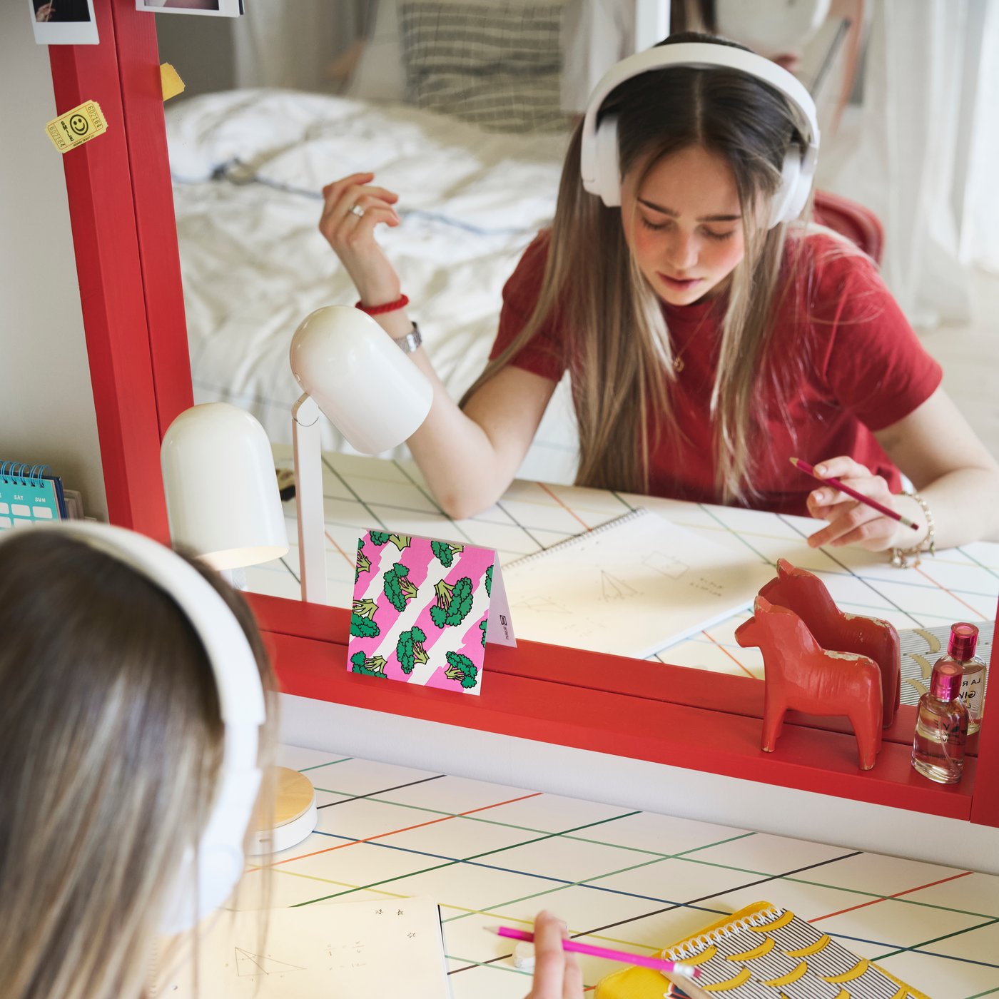 Un'adolescente studia seduta a una scrivania LAGKAPTEN/SPÄND dal motivo a griglia colorato, posizionata di fronte a un grande specchio rosso.