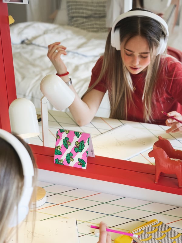 A teenager sitting studying by a LAGKAPTEN/SPÄND desk with a colourful grid pattern placed in front of a big red mirror.