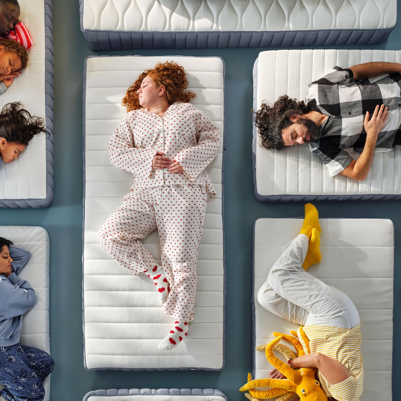 A photo from above of nine people of different ages sleeping on a number of mattresses placed on a gray studio floor.