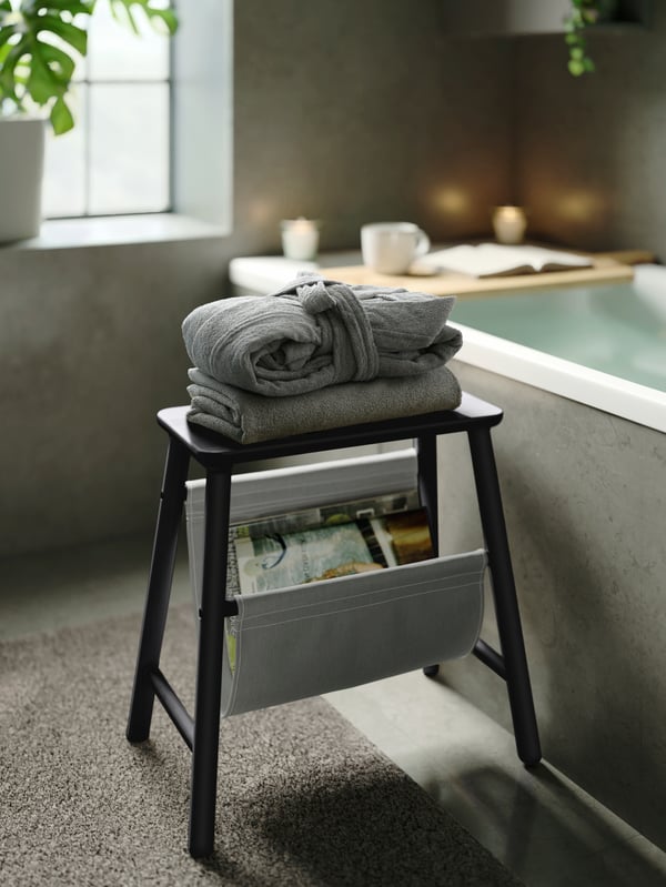 Peignoir ROCKÅN gris bien plié et placé sur un drap de bain gris sur un tabouret en bois situé à côté d’une baignoire avec une étagère pour baignoire posée dessus.