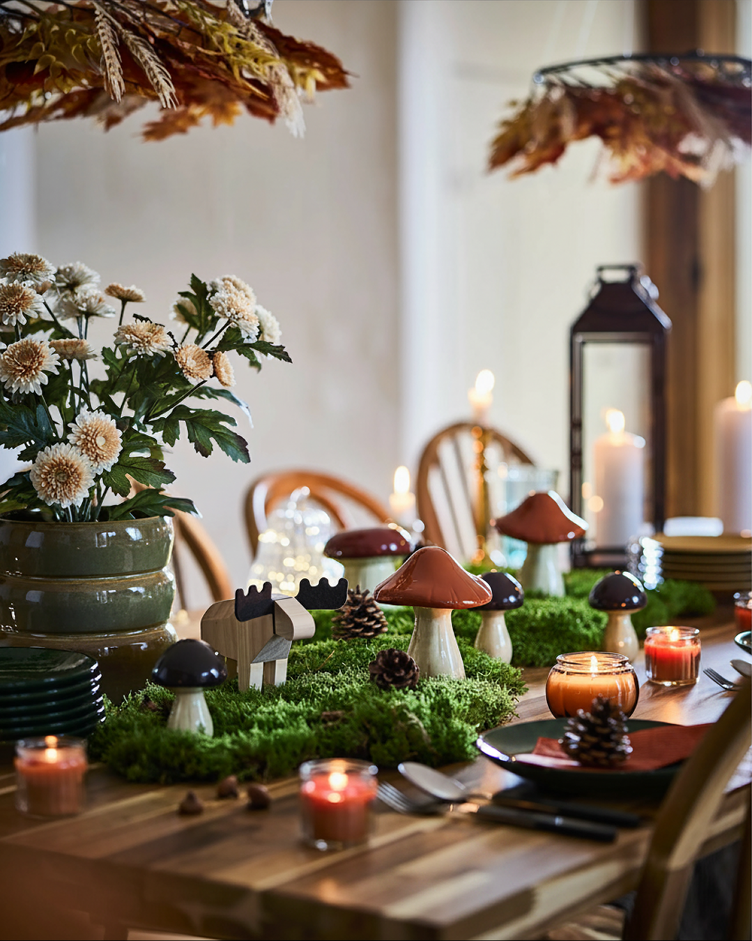 A woodland-themed dining table is set with HÖSTAGILLE decorative mushrooms and moose, lit candles, moss and pinecones.