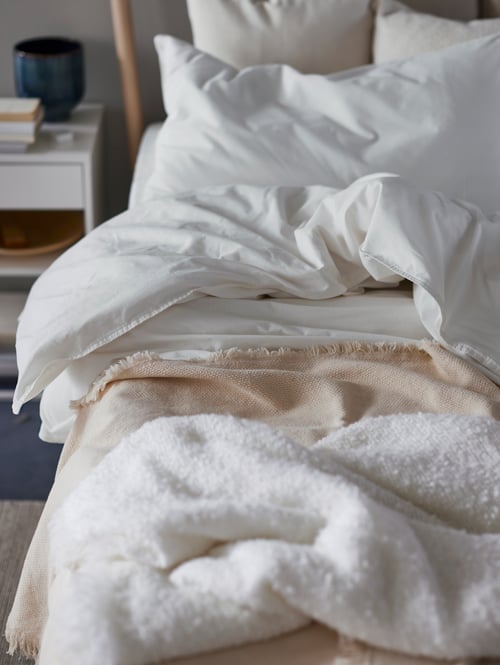 Un cadre de lit en bouleau fait avec du linge de lit blanc ÄNGSLILJA et un plaid beige à côté d'une table de chevet blanche avec une pile de livres.