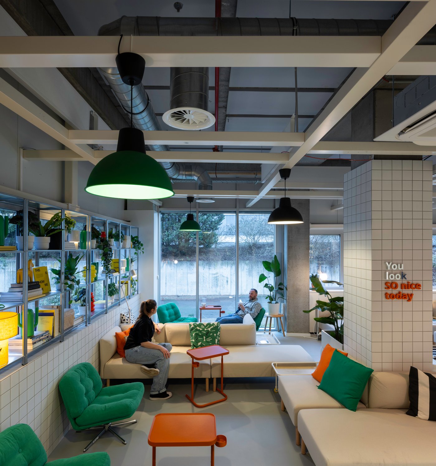 Un espace assis ouvert avec des touches IKEA vertes et orange, entouré de plantes et de sièges aux tons clairs, crée un endroit accueillant pour se retrouver de manière informelle.