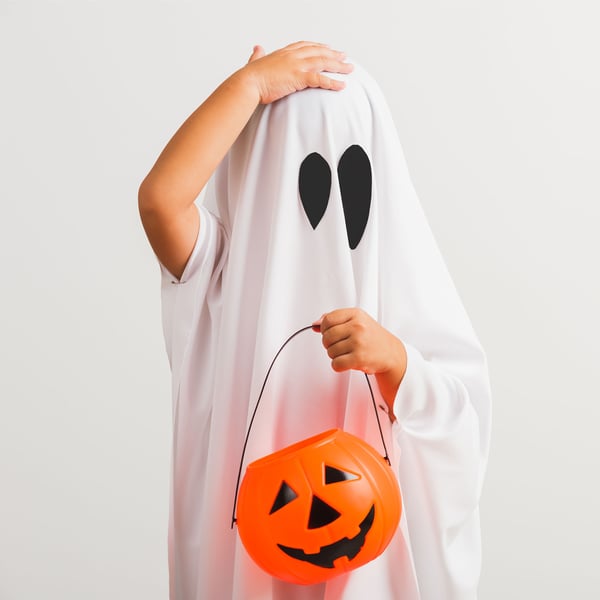 A child wearing a ghost costume and holding a plastic pumpkin basket.