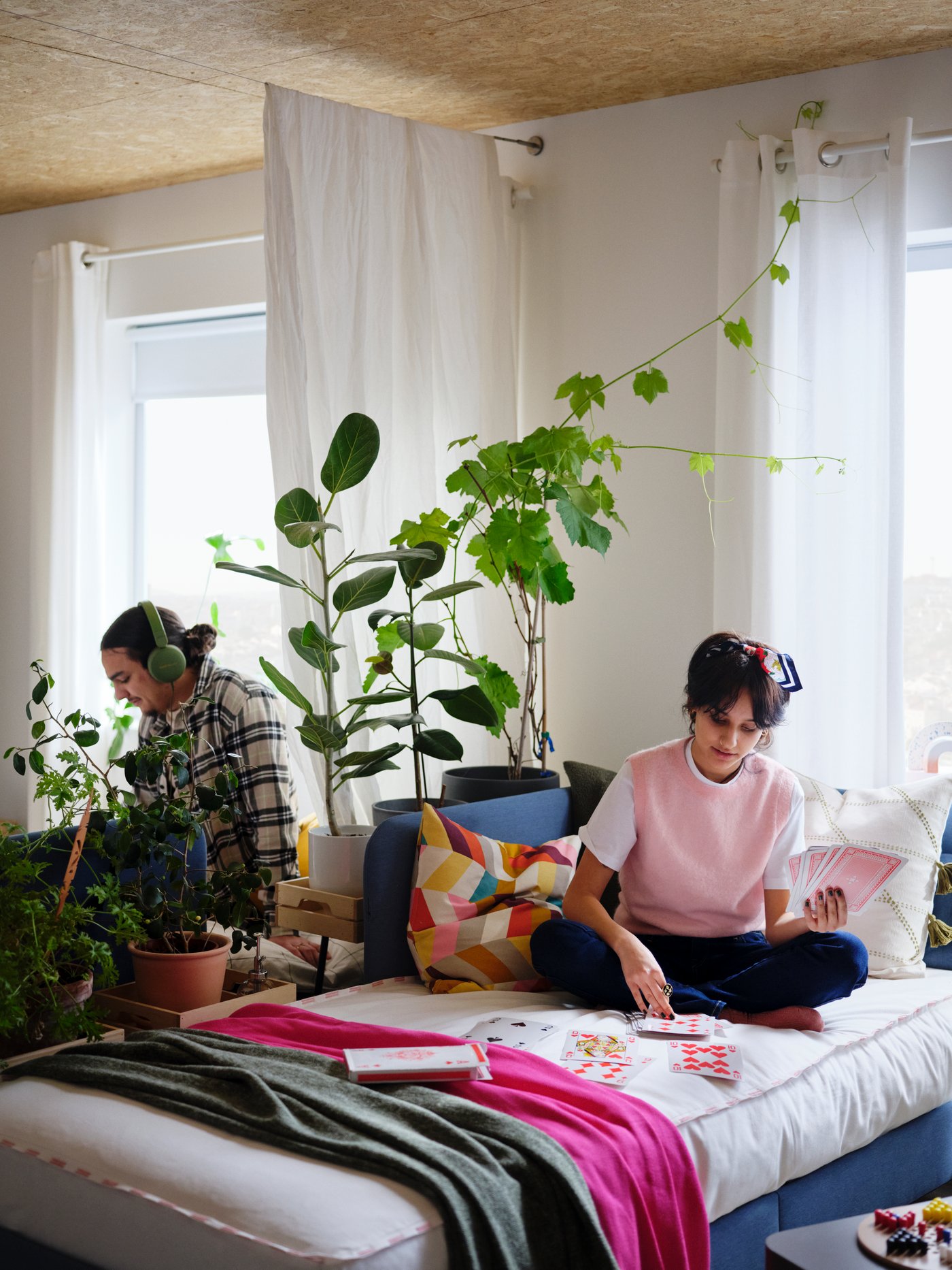 A woman sits on a medium-blue BLÅKULLEN bed. Behind a room-dividing combo of a plant swath and a curtain sits a man.