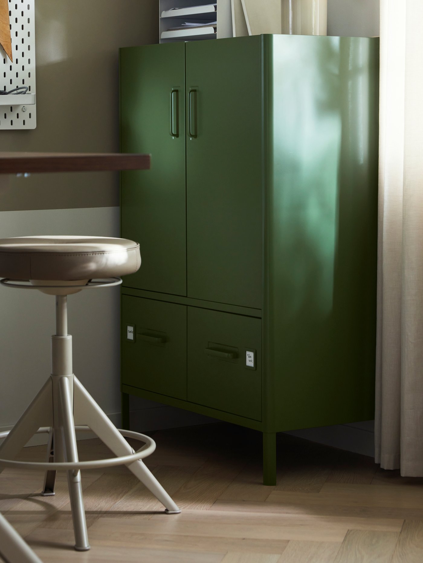 A dark green IDÅSEN cabinet with doors and drawers stands in an office space, with a TROLLBERGET active sit/stand support.