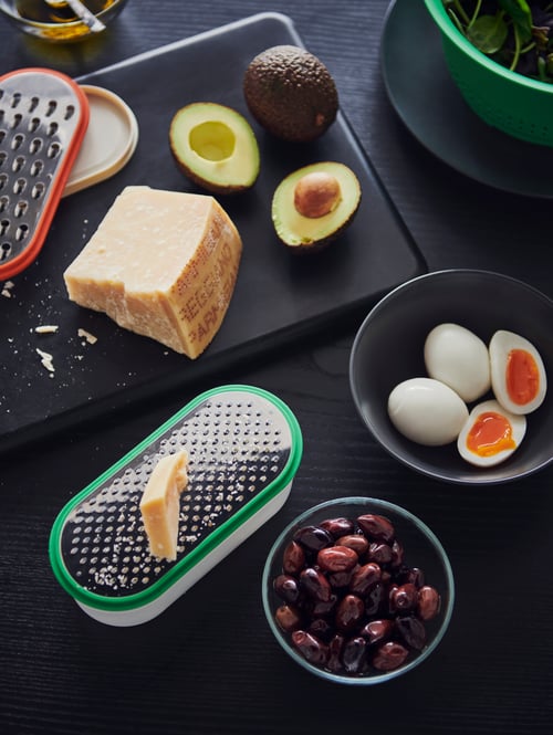 Gros plan sur un comptoir avec une planche à découper, des bols, des ingrédients, et un ensemble de deux râpes et deux contenants en plastique.
