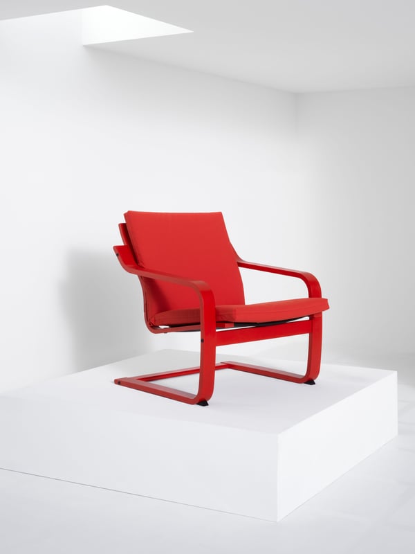 A POÄNG low-back armchair in red stained birch veneer with a red cushion stands on a white podium in a white room.