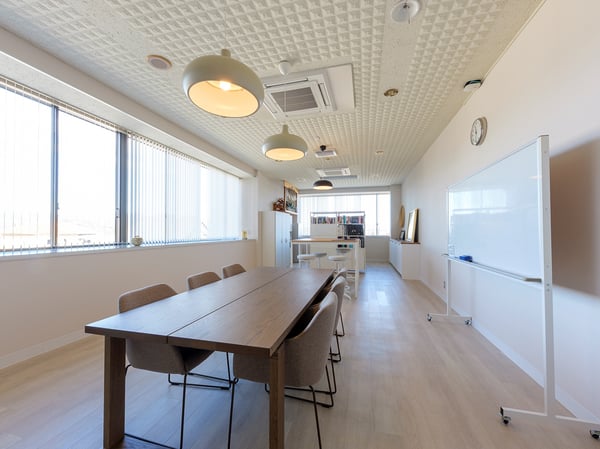 大きな窓から光が入る会議スペース。木製の長テーブルと椅子が並び、壁際にホワイトボードと時計が掛かっている。