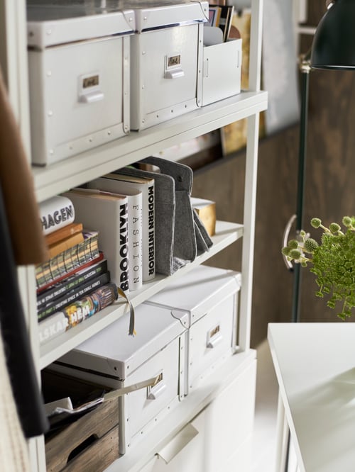 Workspace with white open shelving unit, white storage boxes with lids, books, wooden boxes, green houseplant, pendant lamp.