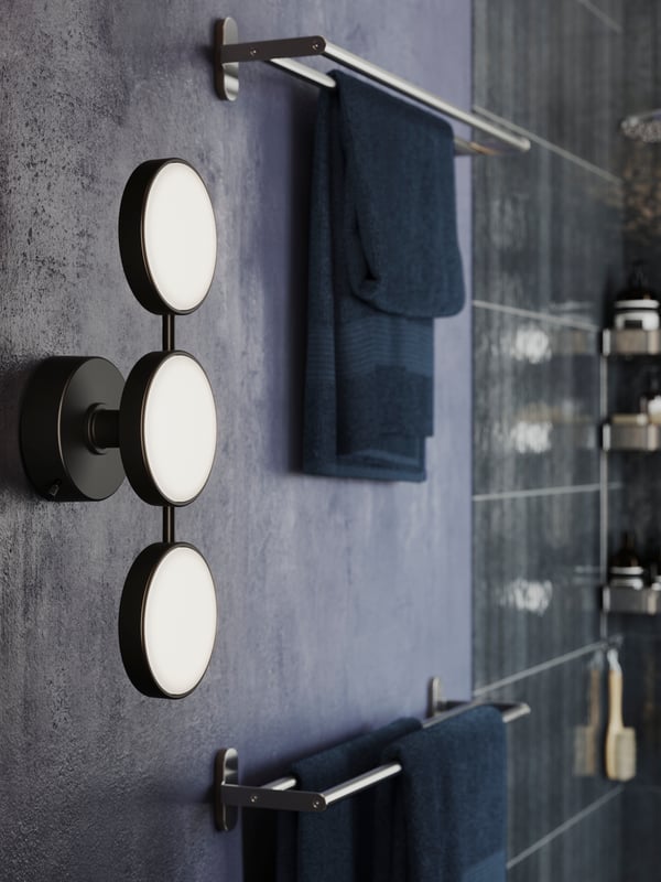 A matte black KABOMBA LED wall lamp is mounted on a bathroom wall next to two BROGRUND towel rails of stainless steel.