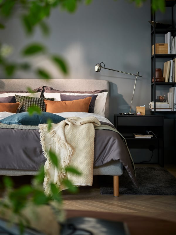 A chrome-plated MODERMOLN work lamp on a black VIKHAMMER bedside table by a beige/grey TÄRNKULLEN upholstered bed.