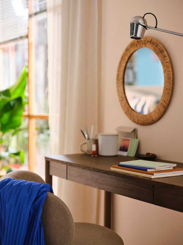 A brown TONSTAD desk against a wall with a rattan KAFFESTRÄD mirror on it. A chrome MODERMOLN work lamp is beside it.