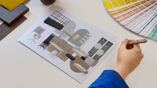 Projektantka wnętrz IKEA prezentująca kartki z wydrukiem kolażu produktowego zaprojektowanego pomieszczeń biznesowych. Obok na blacie leży wzornik kolorów i próbki materiałów.