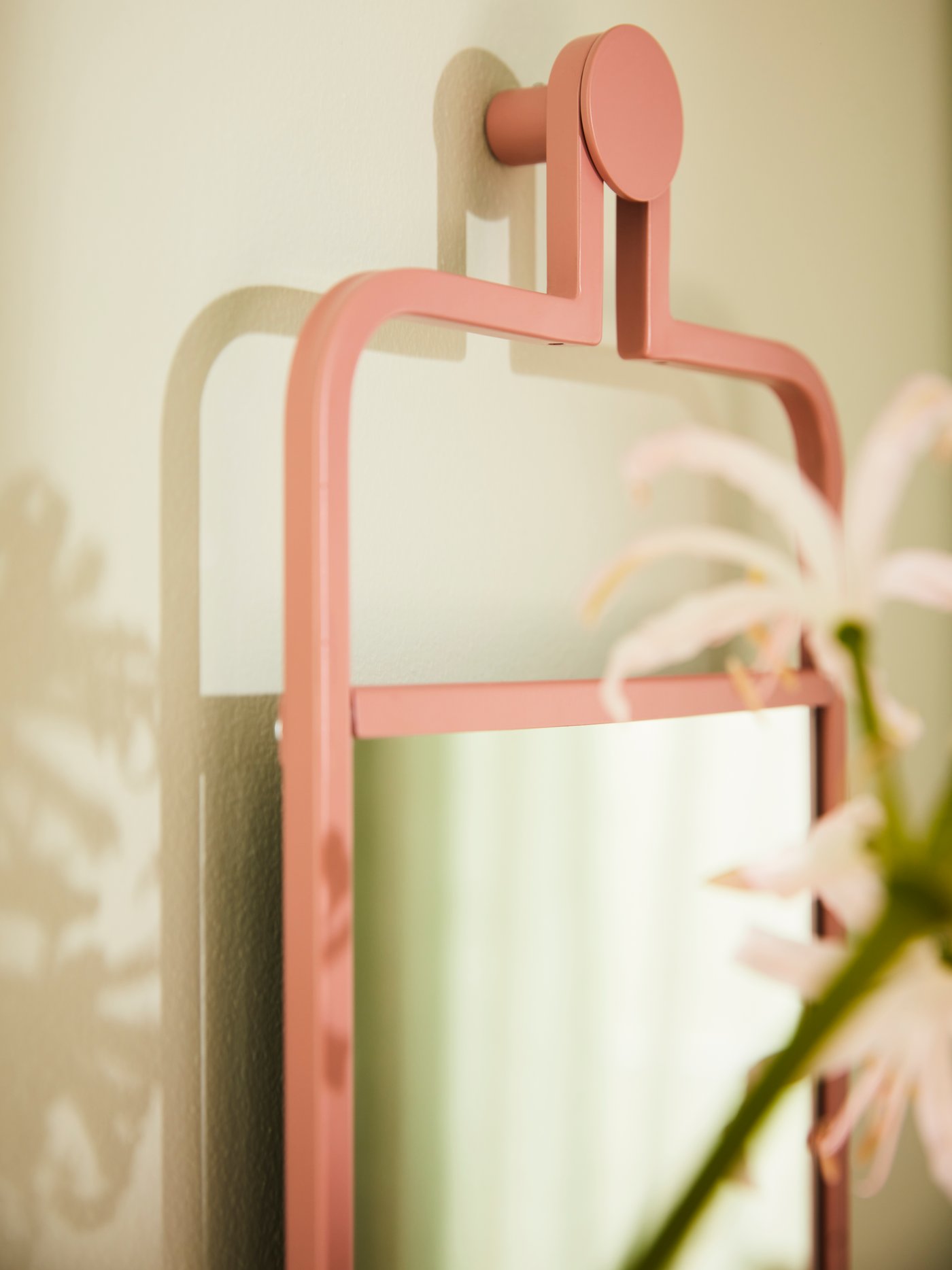 A pink GRANVÅG face mirror hangs on a cream-coloured wall, making a shadow behind it; lilies are blurred in the foreground.