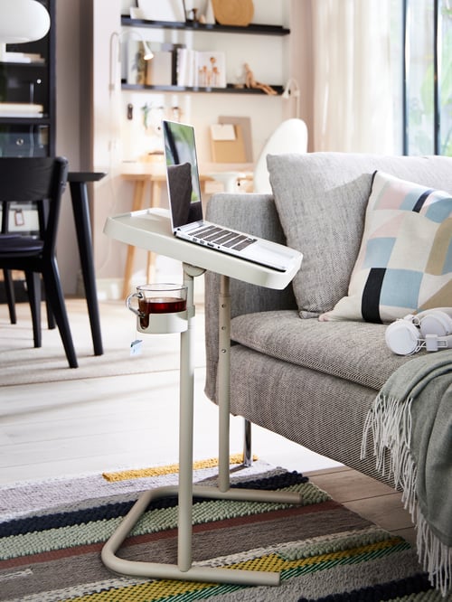 The corner of a beige sofa with a beige BJÖRKÅSEN laptop stand in front of it holding a laptop and a glass mug of tea.