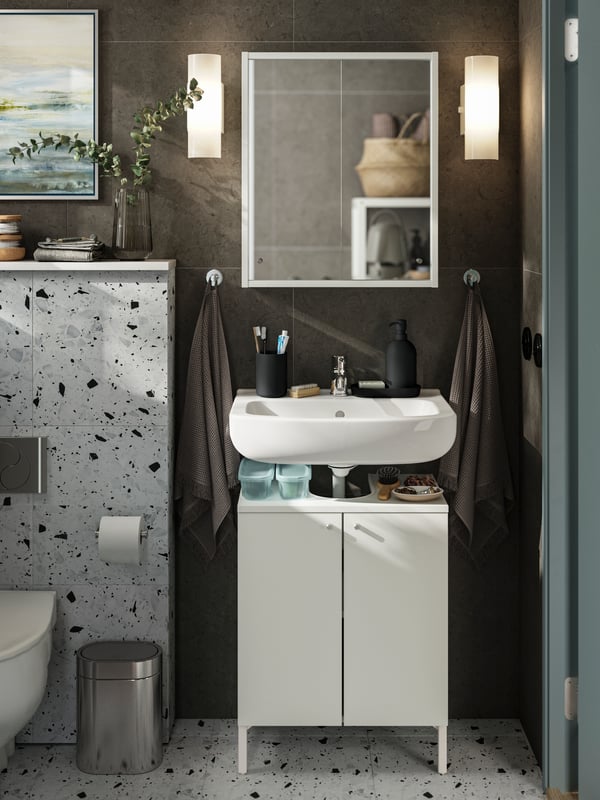 Un élément lavabo NYSJÖN à deux portes sous un lavabo blanc avec des accessoires gris-bleu clair sur le dessus et un miroir sur le mur.