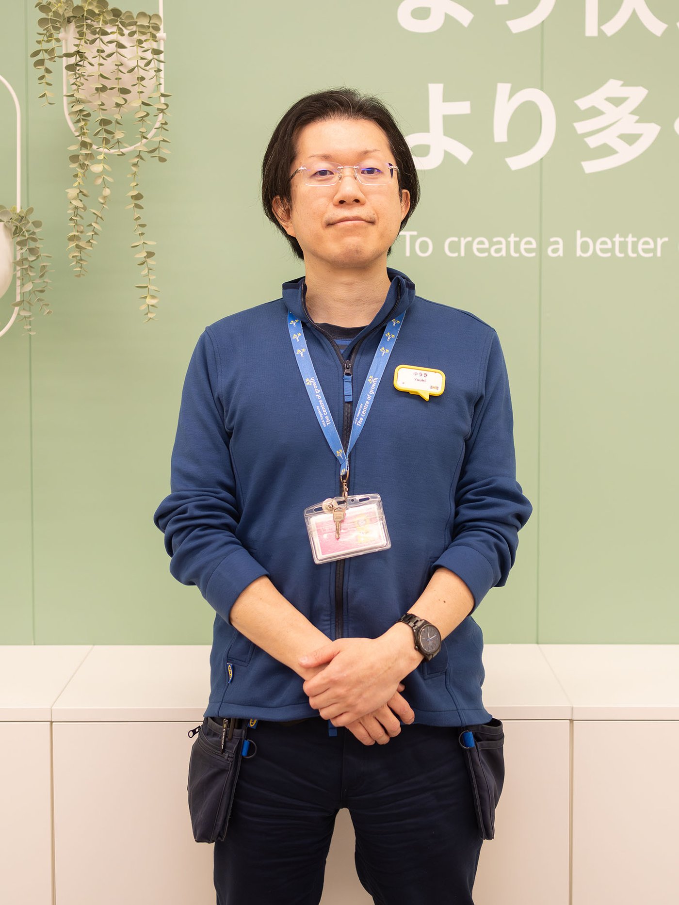 グリーンの壁面とメッセージボードを背景に、青いユニフォームを着たイケアのコワーカーが立っている