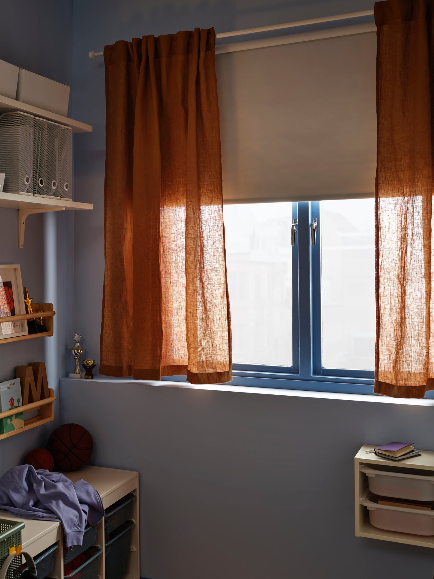 Una persiana de bloqueo de luz opaco FRIDANS blanco en una ventana con el marco azul y un par de cortinas DYTÅG en beige oscuro delante.