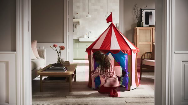 A girl closing the opening to a CIRKUSTÄLT children’s tent on a beige BRÖNDEN rug beside a LISTERBY oak veneer coffee table.