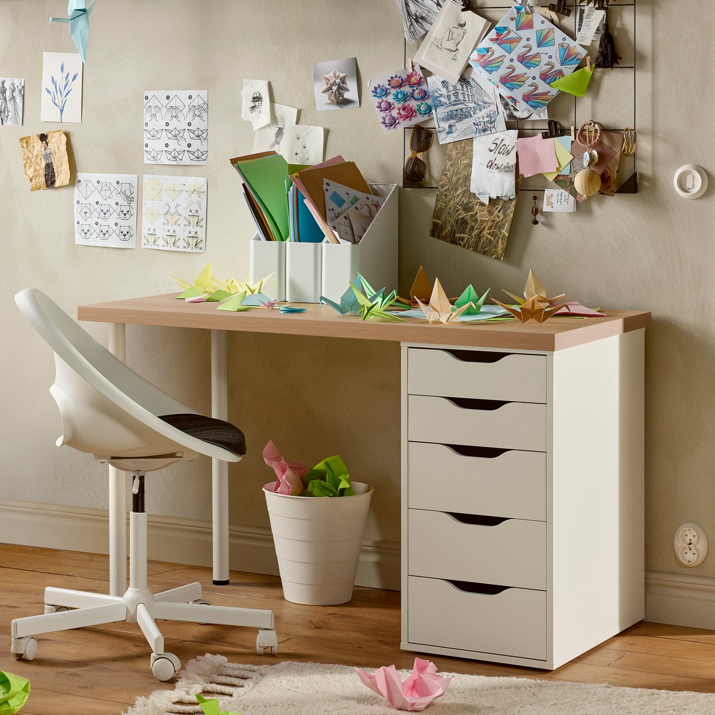 Sedia girevole MALSKÄR bianca davanti a una scrivania con cassetti e pannello memo pieno di appunti. Sopra la scrivania origami colorati, porta documenti e un vaso con pianta.