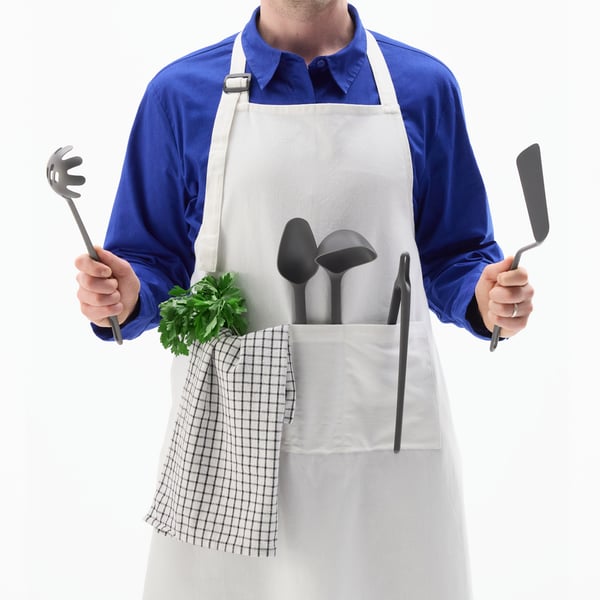 A person in a white apron is holding five large FULLÄNDAD kitchen utensils: turner, spoon, ladle, pasta server and tweezers.