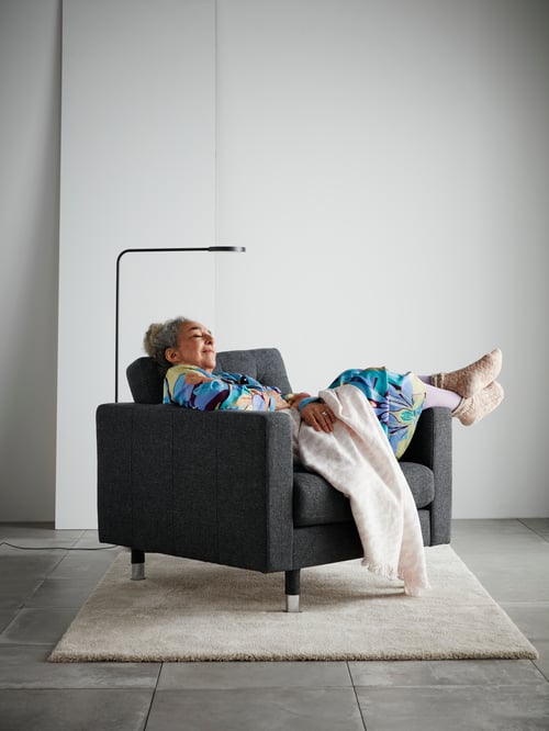 Woman lounging on a dark grey armchair with her feet up, in an empty room with black floor lamp and a white and pink throw.