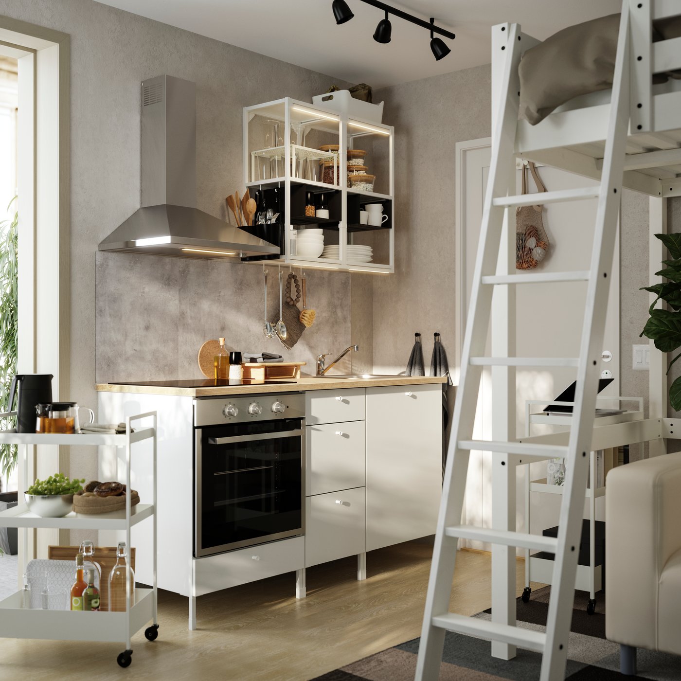 Kompakte IKEA ENHET Singleküche mit weißen Unterschränken und Holzarbeitsplatte, offenem Hängeregal mit Geschirr, Edelstahl-Dunstabzugshaube und Servierwagen; rechts daneben führt eine weiße Leiter zu einem Hochbett im kleinen Wohnraum.