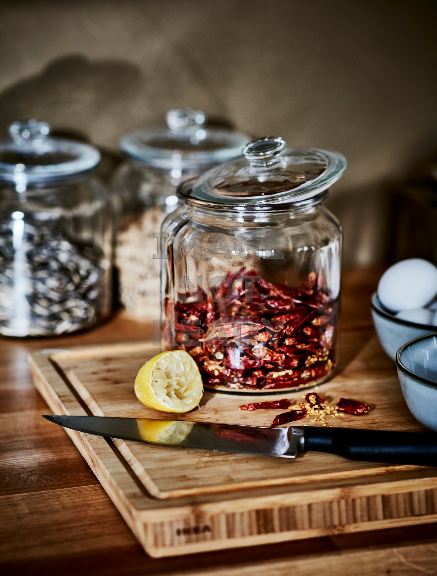 Ein Schneidebrett aus Holz, auf dem ein Messer und eine Zitronenhälfte zu sehen sind. Dahinter steht ein Glas mit roten Chilis.