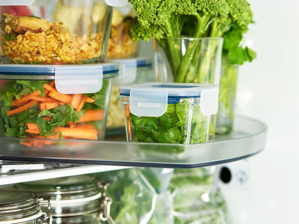 An open refrigerator stocked with various food items. Visible are clear containers with blue lids, one containing a mixed salad, another with what appears to be a grain-based dish, and a third holding cut vegetables including carrots and broccoli. There is also a glass jar filled with a red sauce or preserve. The focus on fresh and prepared foods suggests an emphasis on healthy eating or meal preparation.