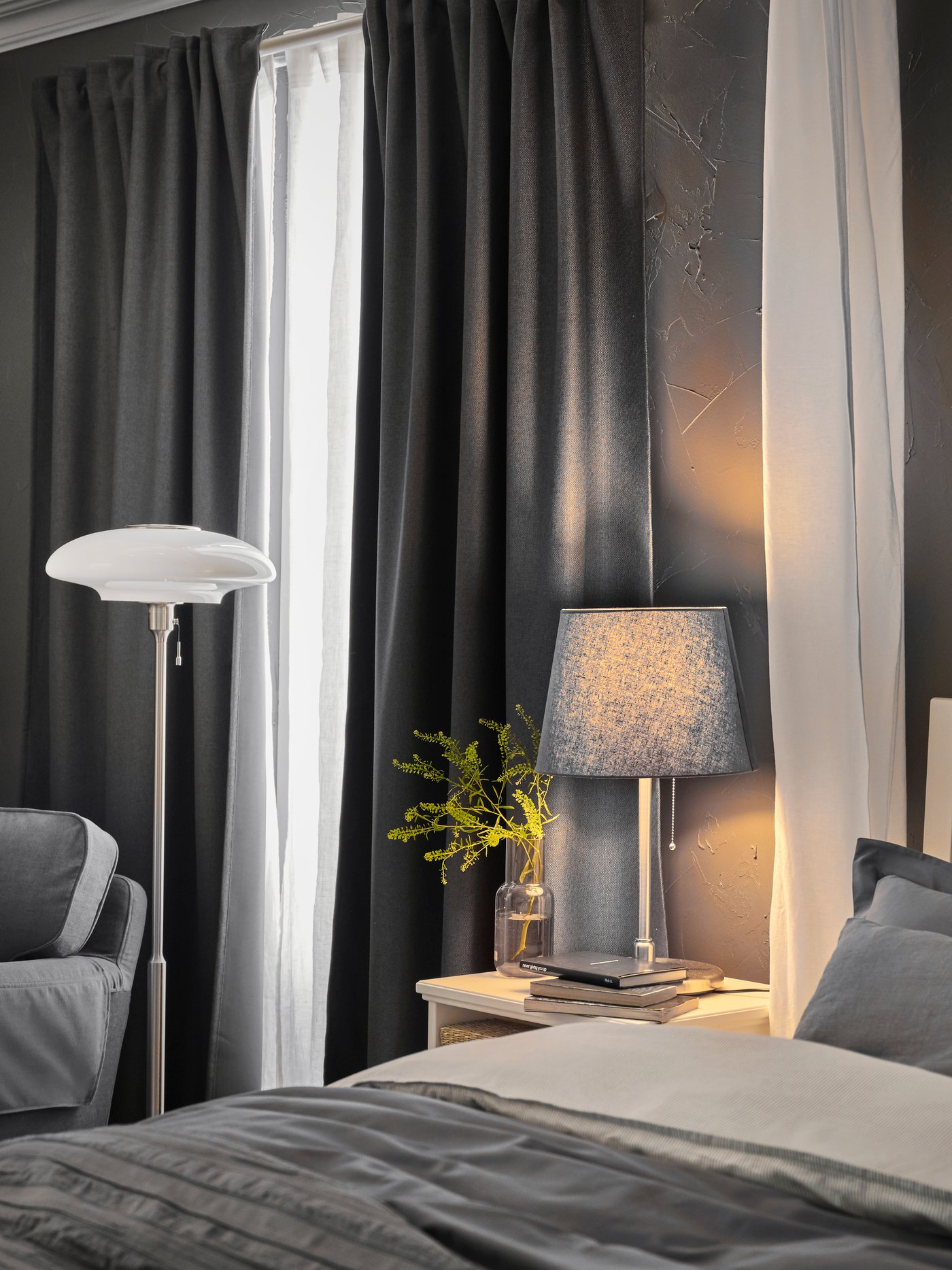 A white curtain hangs down at the side of an IDANÄS bed near a TÄLLBYN floor lamp and a table lamp on a bedside table.