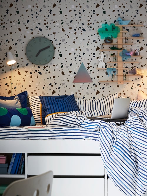 White SLÄKT bed in a room with speckled wallpaper, a clock and shelves on the wall, striped bed linen and coloured pillows.