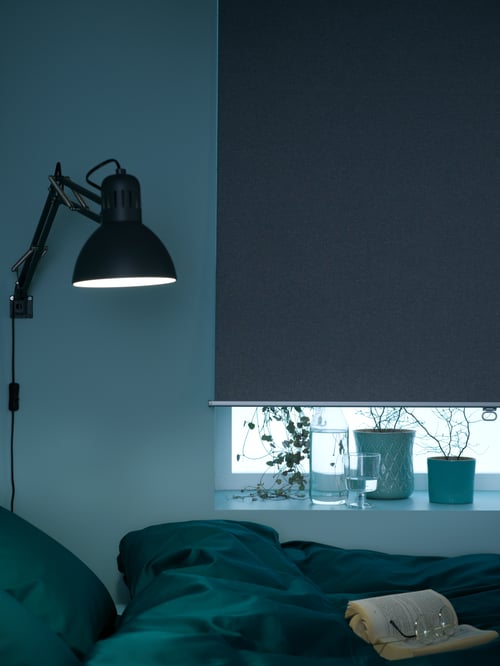 Chambre sombre avec un store à enrouleur occultant TRETUR tiré presque jusqu’en bas, avec quelques cache-pots posés sur le rebord de la fenêtre.