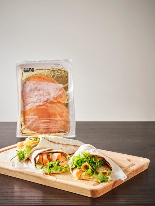 An unopened packet of SJÖRAPPORT cold-smoked salmon is behind a chopping board with bread wraps containing salmon and salad.