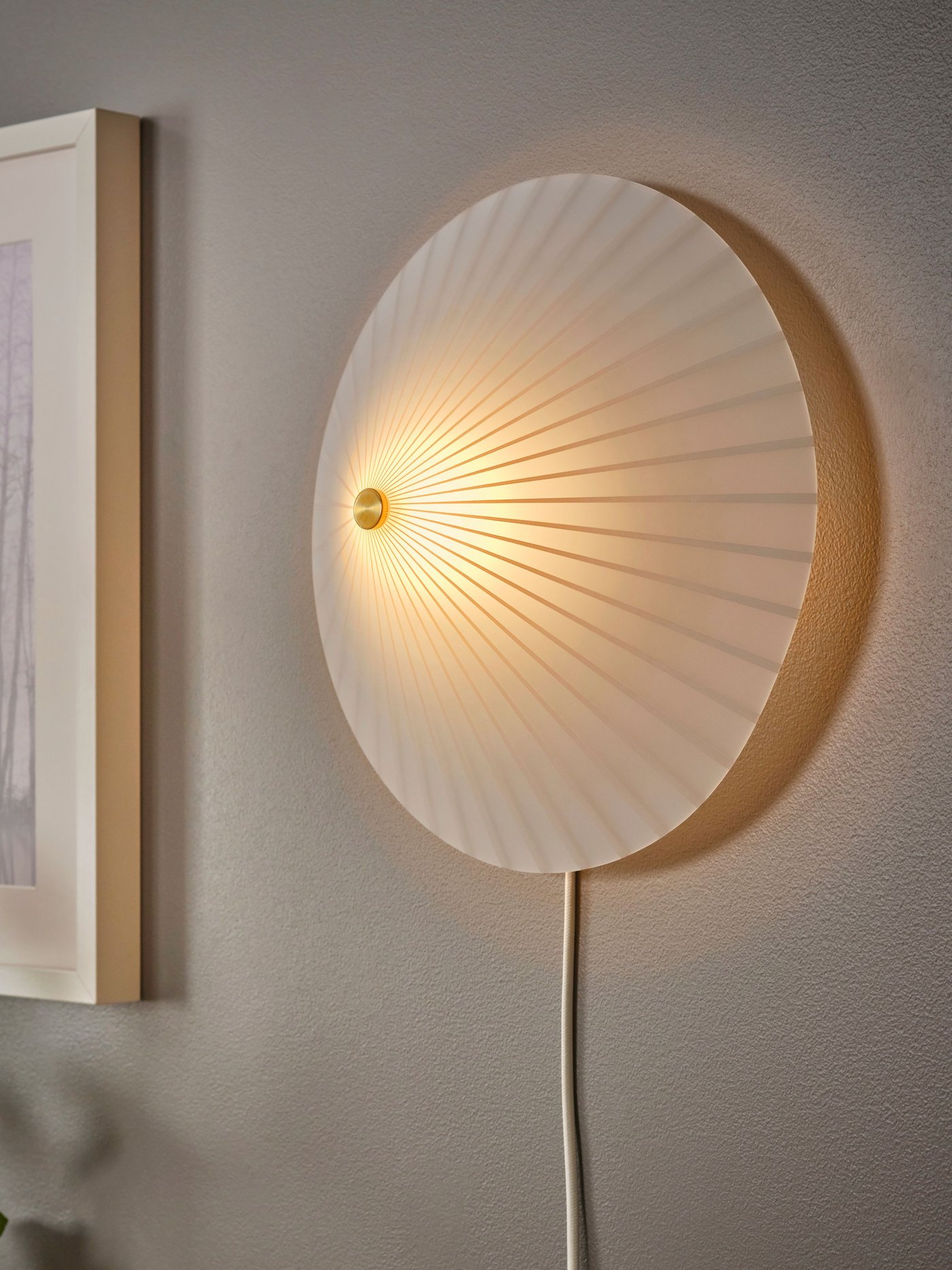 En RORKULT vägglampa i randigt mönster/vitt glas är monterad på en vägg nära en tavla i ram som också sitter på väggen.