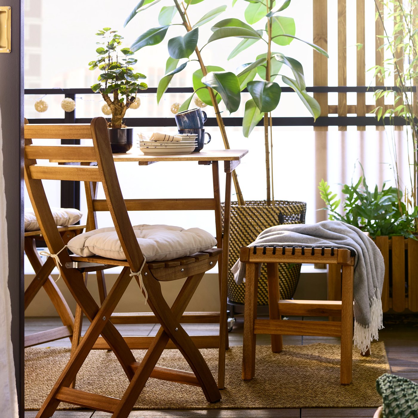 A light brown stained acacia ASKHOLMEN table and foldable chair face the view in a balcony with plants and natural materials.